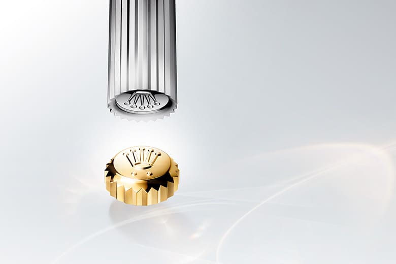 A close-up composition featuring a gold Rolex crown and a steel die against a bright, minimal backdrop.